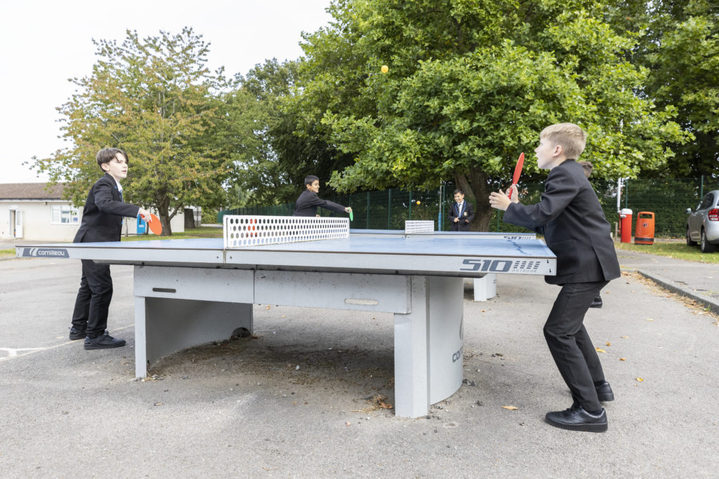 Two male students can be seen standing in the playground, playing Table Tennis together.