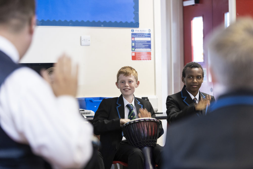 Students are seen playing the drums with their hands during a Music lesson.
