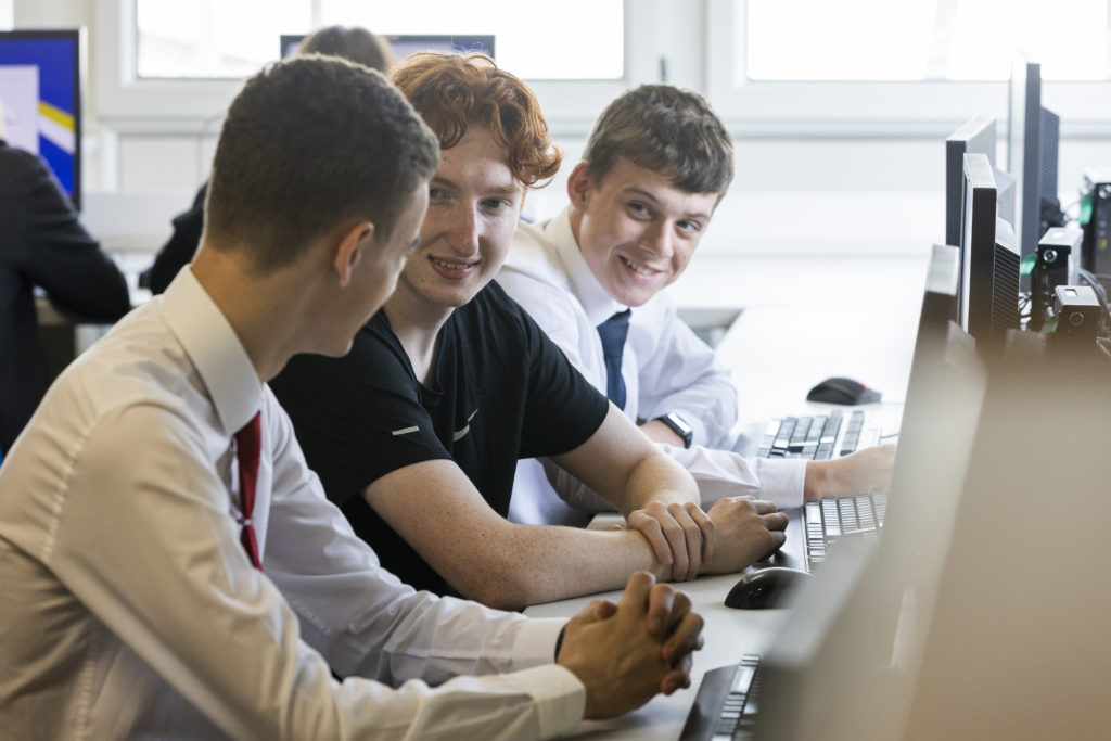 Three male students are sat a desks working on computers. They are collaborating with one another to complete their work.