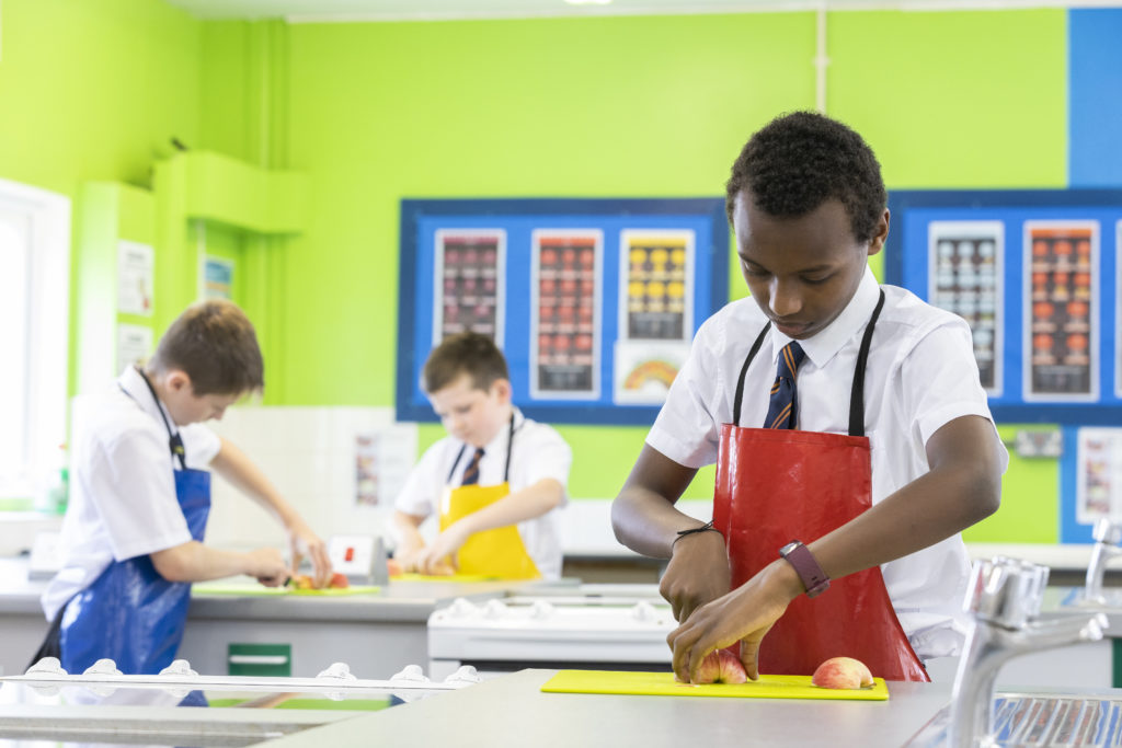 Three students are seen cooking and wearing coloured aprons during a Food Technology class.