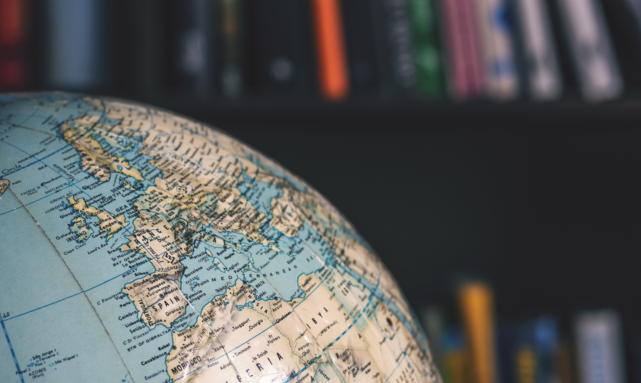 Close-up photo of a globe of the world seen in the foreground with a large shelf full of books in the background.