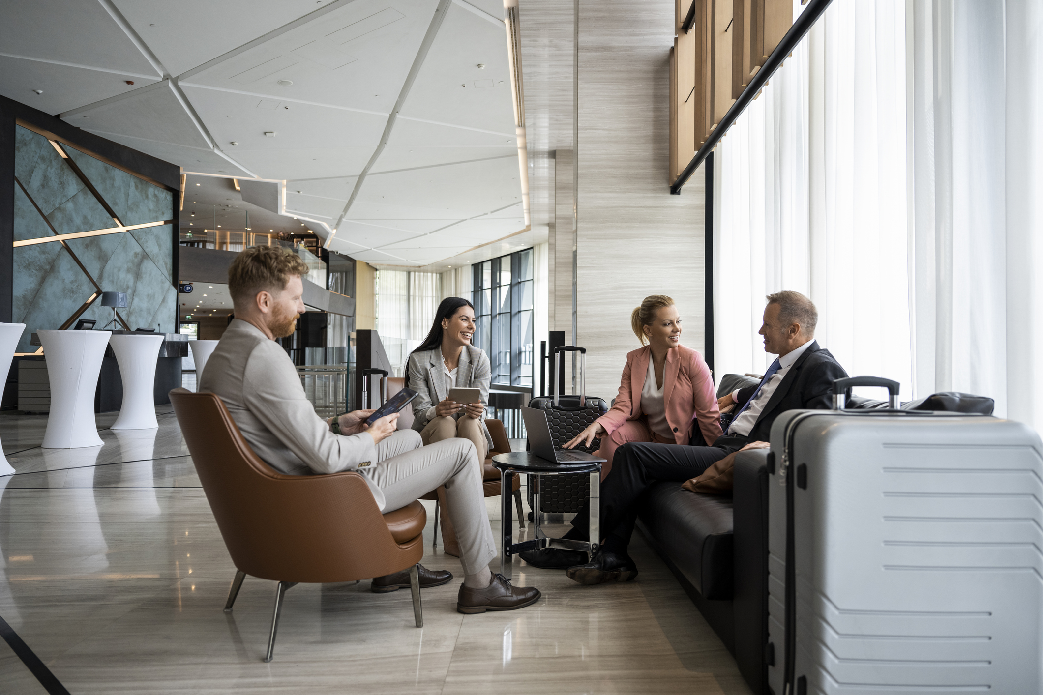 Four colleagues seen sitting in a luxury hotel lobby; talking, smiling and preparing for a business meeting. Two of them are holding a smart device and the other two are using a laptop.