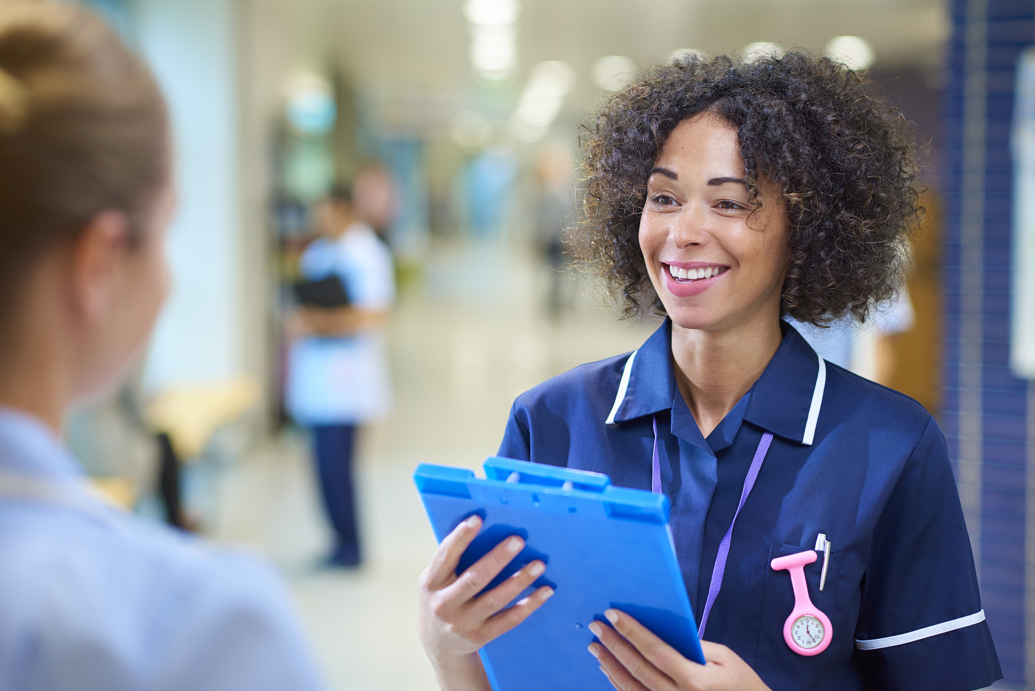 A senior nurse is pictured smiling at another junior staff member in a hospital corridor. The senior nurse can be seen grasping a blue clipboard in her hands.