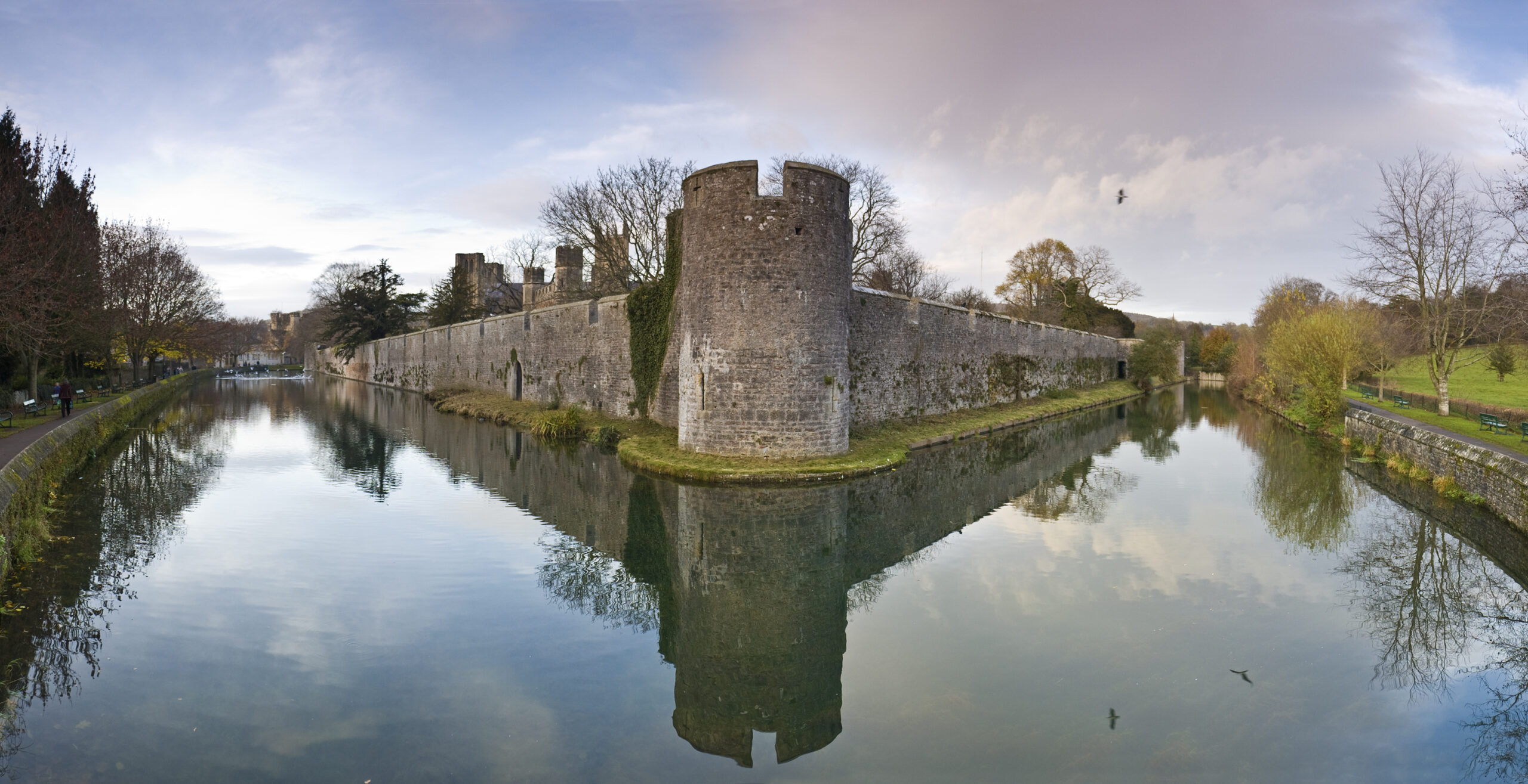 Still waters of a wide moat reflecting the historic stone walls and battlements of the Bishop's Palace at Wells, Somerset, UK.