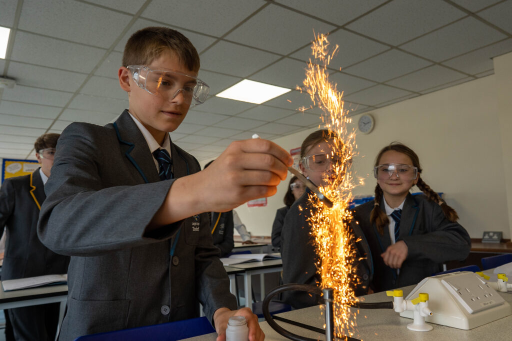 Three students, a boy and two girls, are pictured participating in a Science experiment together, using a Bunsen Burner. Peers behind them can be seen looking on in curiosity.