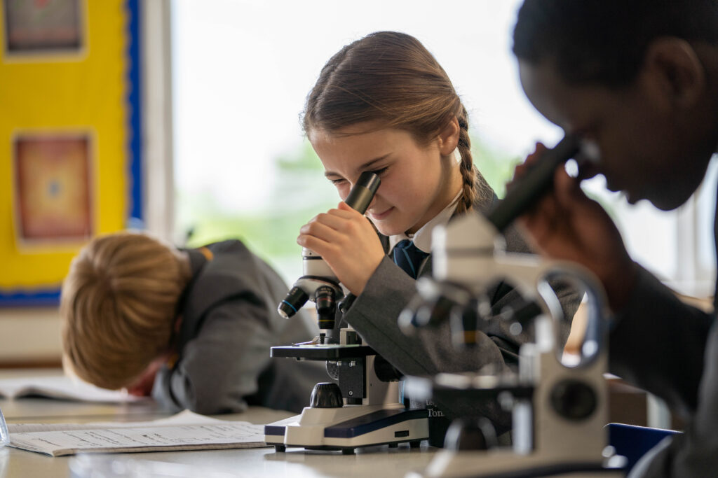 A female student can be seen dressed in her academy uniform, whilst conducting a Science experiment using a microscope in a lab class. She is shown closing one eye and using the other to peer down the lens.