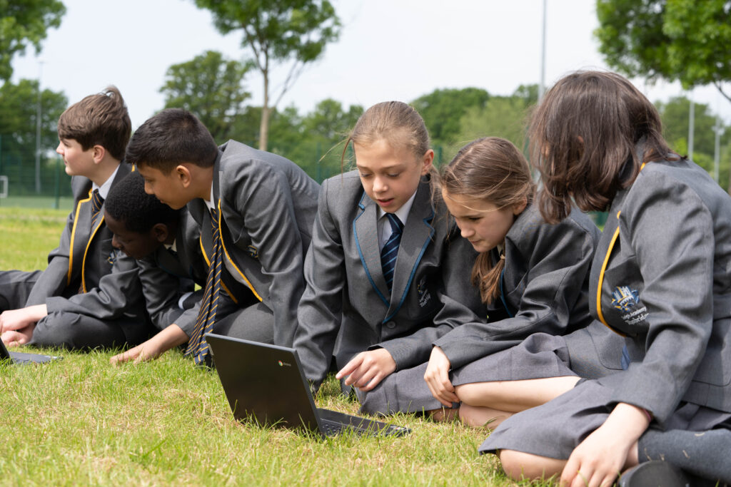 Five students, two boys and three girls, are pictured outdoors on the academy grounds, wearing their academy uniform. They are seen looking at laptop screens, whilst sitting on a grassy area.