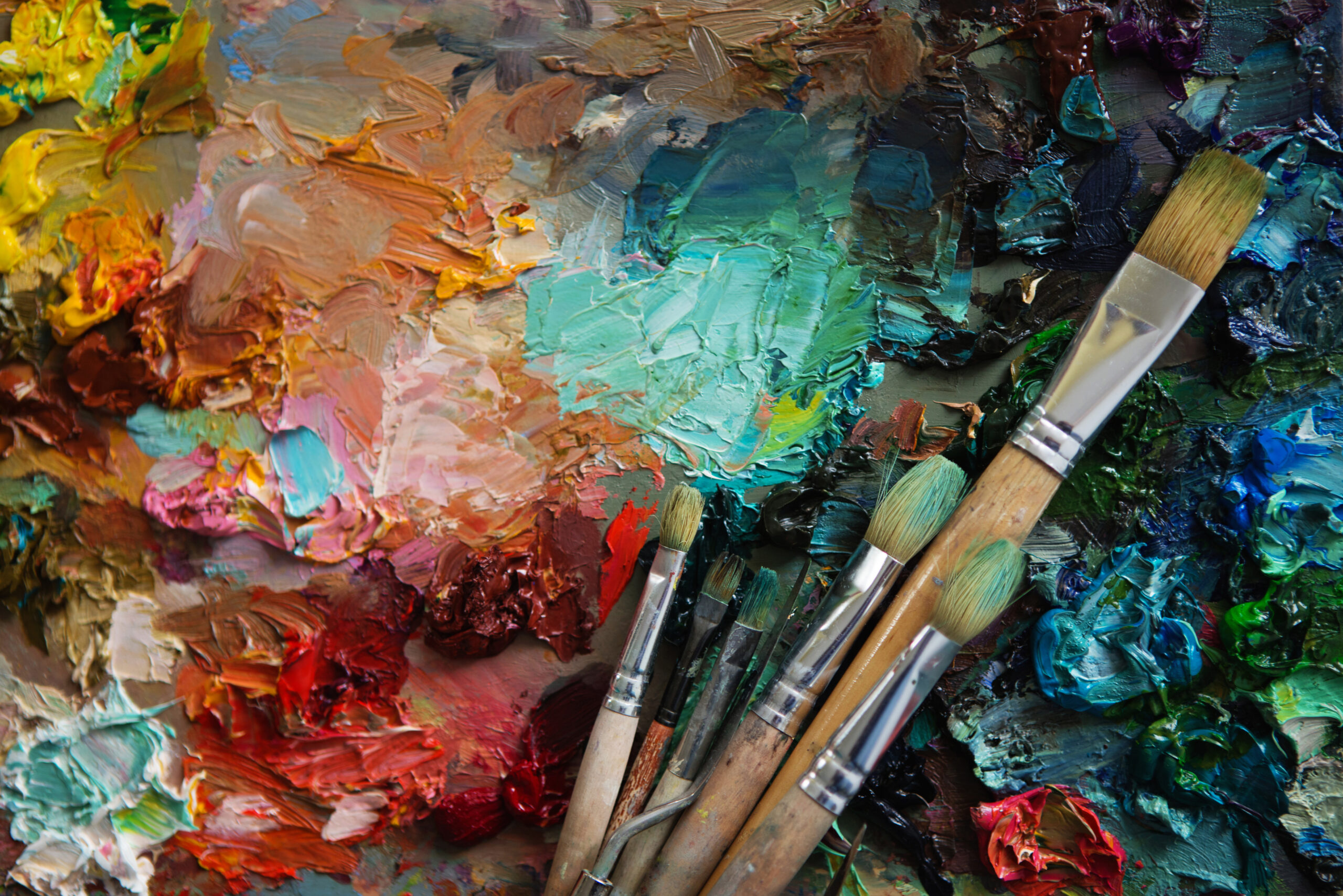A set of paintbrushes are pictured resting on an art canvas where many different paint colours have been used.