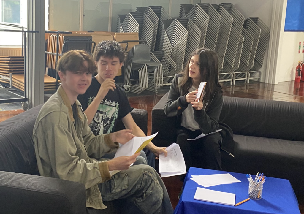 A group of three students seen sat down on sofas in an open area of the academy building, smiling as they open their results envelopes.