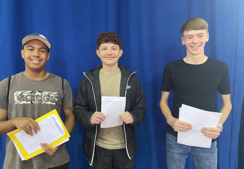 Three male students are pictured standing alongside one another, smiling as they open their results envelopes.