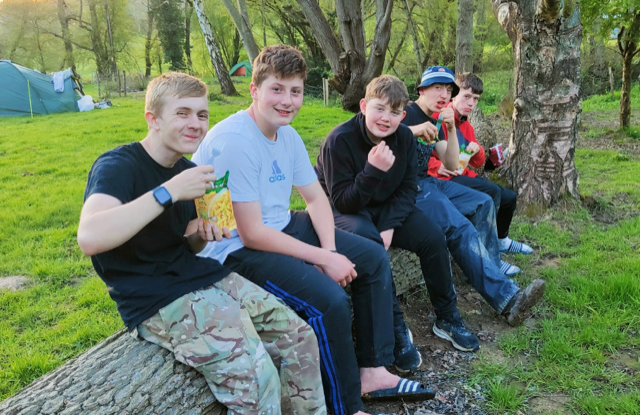 A group of five students seen sat along a log together, smiling for a photo whilst eating.