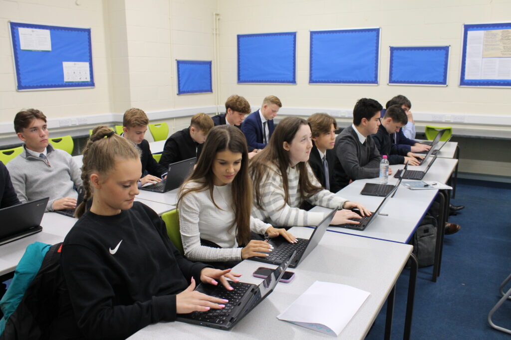 A classroom is pictured with a large group of students sat at its desks. Each student is seen using a laptop computer to assist them with their learning, as they pay attention to the teacher at the front of the room.
