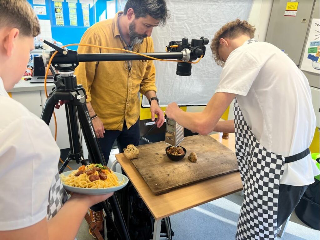 2 students preparing food, supervised by Rob Morris, with a camera on a tripod pointed at the dish on the table